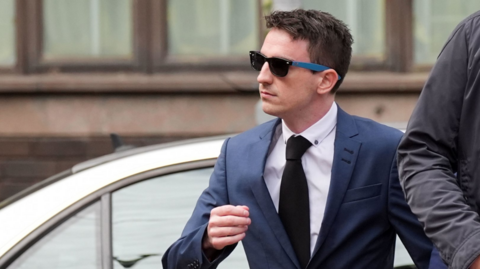 A man with dark hair wears sunglasses, a blue suit, and a black tie, and looks to the left, while standing in front of a silver car