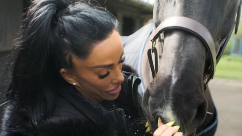 Katie Price is pictured up close with a horse. She has her dark hair tied up in a ponytail.