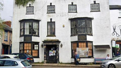 The derelict frontage of a hotel building. There are three storeys, and the building is painted white. There are bay windows on the ground and first floors. There are a number of cars parked in front of the building.