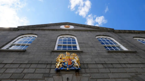 The picture shows the exterior of a formal government or judicial building made of grey stone. The architecture features three large arched windows and a triangular pediment at the top. There are two prominent crests displayed:
One smaller crest is positioned high up in the centre of the pediment.
A larger, ornate coat of arms is mounted below the windows. This coat of arms includes a crowned lion and a unicorn.