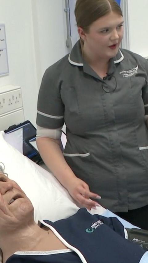A lifelike patient mannequin on a bed. There is a woman in grey nursing uniform next to the bed.