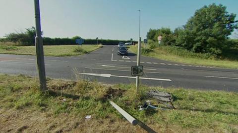 A road junction with cars approaching. In the foreground you can see a post that has been knocked over and some debris next to a speed camera sign. There are grass verges on all sides of the junction and trees to the right. Cars waiting at the junction can be seen in the background.