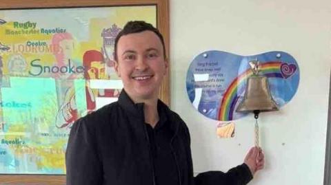 Man with dark brown short hair wears an all black outfit and is smiling - Sam Birchall. The 24-year-old stands in a hospital room in front of a bronze bell on a rainbow mount. He holds the string of the bell as if to begin ringing it.