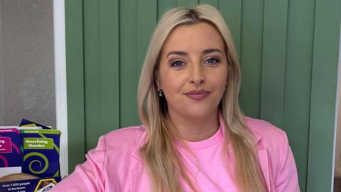 Órlaithí Flynn has blonde hair, a side parting and gold earrings. She is wearing a 
bright pink t shirt and a pink jacket and is standing in front of a wood-panelled wall which is painted in green