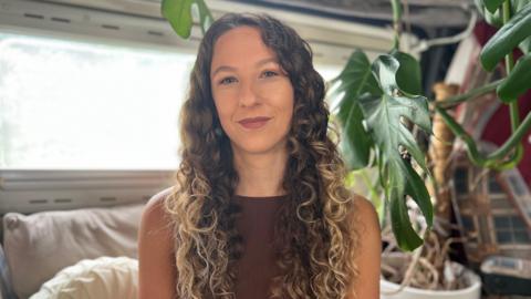 Sophie Robert pictured with long, curly brunette hair that turns blonde towards the end sitting indoors beside a large leafy houseplant, wearing a sleeveless brown top.
