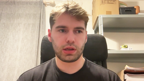 Olly Foster, who was one of the passengers on board the train. He is white, has brown/blonde hair and stubble. He is wearing a black t shirt and can be seen from top of his chest up. He is speaking to BBC news and this image is a still taken from that live video interview. He appears to be sitting in a computer chair - possibly in a spare room of a house. There are shelves in the background and the grey curtains behind him are closed.