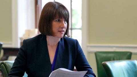 Rachel Reeves sits at a desk with empty chairs beside her. She's leaning forward and appears to be listening and is holding a piece of A4 paper