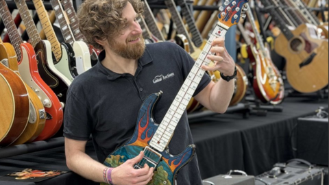 A man with brown curly hair and beard smiling and holding a blue guitar with orange flames on its body and a thermometer up its stem - with a rack of guitars in the background.