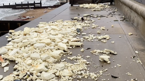 A section of concrete on top of a sea wall is covered with piles of light yellow insulation, from tiny pieces to sections about a foot long. Blackened bananas can also be seen.