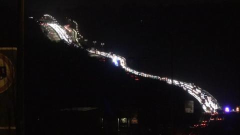 A night time scene showing a long bendy road with a queue of traffic, with white car headlights lined up. On the right edge of the picture blue flashing lights can just be seen.