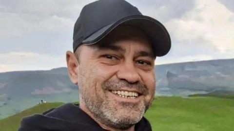 A man in his early 40s, wearing a baseball cap and smiling into the camera. He has grey stubble and is standing against the backdrop of some green hills.