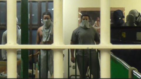 A group of prisoners are wearing grey clothes and are standing in front of a wall. They have face coverings on and are behind bars.