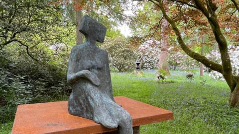 A grey statue of a faceless person with a square head, sat on a terracotta ledge. The statue is situated in a green garden.