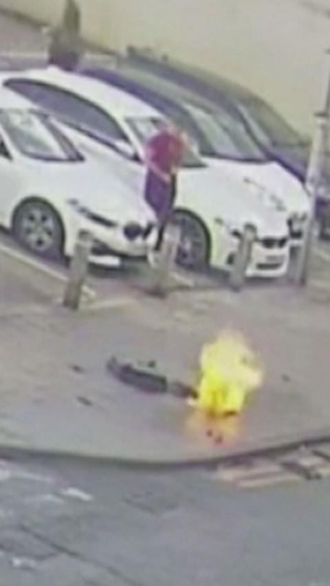 A fire on a pavement on a residential road. Behind it is a car park with several white cars. A fuzzy figure is next to them.