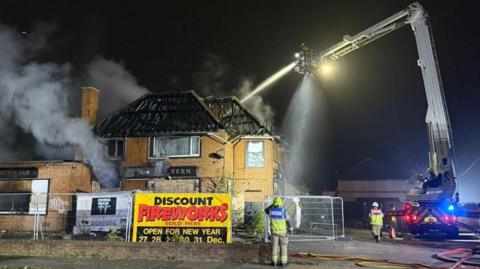 Fire officers standing near a burning building. Smoke is pouring out of a window and the roof is a shell. A fire officer is on a tall platform and is using a hose to pour water on the building from the height.