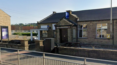 A one storey building with benches outside. Above the door a sign reads: Ryshworth Club