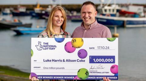 Luke Harris (on the right, a man with short brown hair and smiling looking into the camera, wearing a red and white chequered shirt) and Alison Coke (on the left, a woman with blonde hair looking directly into the camera and smiling, wearing a patterned dress consisting of white, green and red) holding a cheque from the National Lottery for £1m in front of a dock area