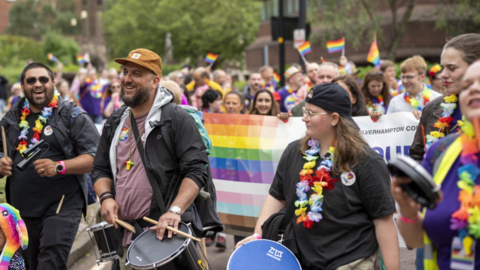A crowd of people marching, with three in the foreground carrying drums and wearing colourful rainbow garlands