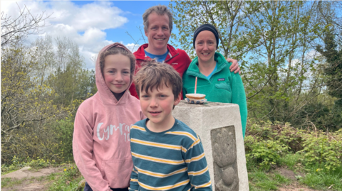 Man, woman and two children stand around a trig pillar. On top of the trig pillar is a small cake with a candle sticking out of the top.