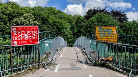 Baths Bridge which is a curved concrete path with green metal railings on both sides. There are silver temporary barriers lining the bridge in front of the railings and two large signs at the beginning of the bridge. The one on the left is red and reads 'Cyclists dismount', while the one on the right is yellow and reads: 'Weak bridge please give way to oncoming pedestrians and maintain social distancing regulations. Cyclists must dismount'. They have both been defaced with graffiti. There are white broken up lines signalling cycle lanes on either side of the bridge. Over the bridge there are lots of green trees.