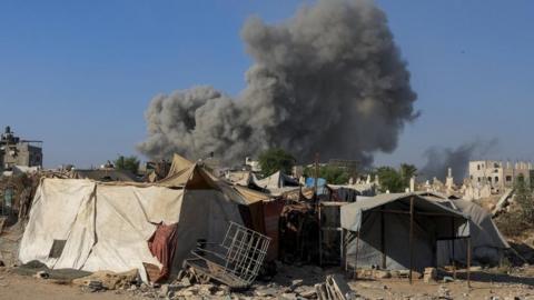 Ramshackle tent-like structures, one has a kind of a metal grid in front, with a large plume of smoke behind them and two half-finished barely-standing building on either side, in Gaza City on 24 August 2025 