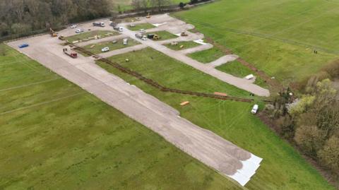 An aerial shot of a green field edged by a country lane along the top. Concrete hard standing has been laid in a grid pattern and a trench has been dug through middle of the site. There are vans, diggers and cars moving around the site. 
