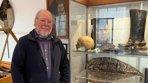 Robert Bell, the museum's curator, is wearing a black fleece and checked shirt and standing next to a glass cabinet. Inside the cabinet are items including a skull, a large boot and a metal engraved item in the shape of an oval.