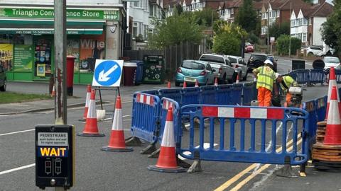 Builders in high vis jackets stand in a road surrounded by royal blue barriers and red and white traffic cones  as they mark out an area of widened pavement using white paint. A convenience store and a number of houses and cars can be seen in the background. 