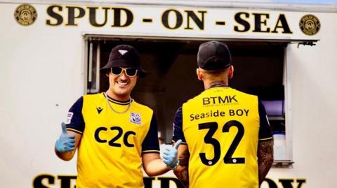Bradley Gibbs and Max Linduschka are standing in front of their Spud-on-Sea van which is white and has the name at the top. Mr Gibbs is on the left looking towards the camera, and Mr Linduschka is on the right with his back towards the camera. They are both wearing a yellow and black T-shirt. 