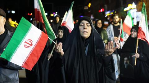 A woman holding an Iranian flag and praying on a protest in support of Iran's supreme leader Mojtaba Khamenei  in Tehran, March 25th