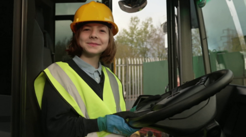 Tommy smiles as he sits behind the wheel of a bin lorry. He has collar length brown hair and wears a blue shirt under a black jumper with a high-vis vest over. He wears a yellow hard hat and blue gloves