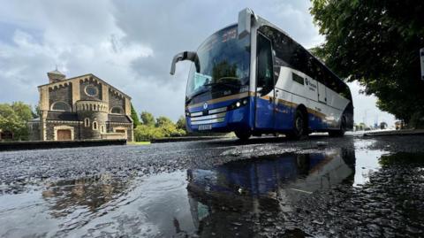 Blue and white Ulsterbus coach, low camera angle of with puddle in foreground and building in background 