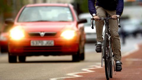 Cyclist in dark blue shirt and khaki trousers in cycle lane close to a red car with headlights on