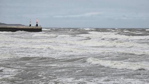 Choppy grey and white waves crash into the shore. An illuminated red light shines from the top of a small lighthouse at the end of a pier.