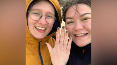 Ella Cook, pictured left, smiles at the camera. She is wearing glasses and a yellow puffer jacket with the hood up. Her fiancé, Kai Poa-Tams, is on her right. Kai smiles at the camera and is showing her engagement ring on her hand.