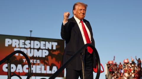 Donald Trump stands with his fist raised, looking defiant. He's wearing a black suit with a red tie, in front of a sign which reads 'President Trump takes Coachella'