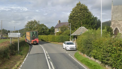 The A591 road is a singe carriageway at Chapel near Bassenthwaite. There is a farm truck travelling one way and a white car parked outside a church on the right. There are green bushes lining the road.