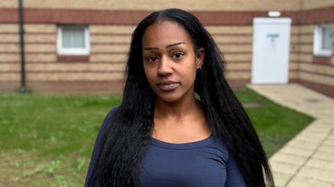 The head and shoulders of a woman with long black hair and a dark blue top standing outside a bricked building.