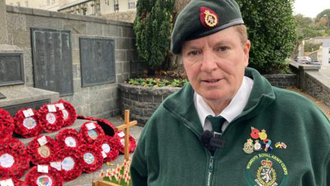 Ex-Army veteran Linda Garnham from the Women's Royal Army Corps Association dressed in bottle green-coloured uniform, complete with badges, beret, tie and beret - stood next to wreaths of poppies, beside the war memorial in St Peter Port, Guernsey.