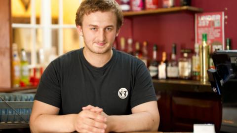 Alex Barry, a young man with blond hair, stands behind an old fashioned bar in a pub.