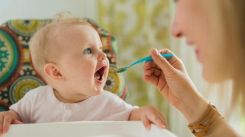 A stock image of a baby with blue eyes being spoon fed by their mother.