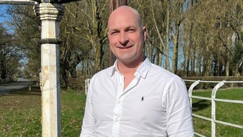 Anthony Harrrison, a man wearing a white shirt and stood outdoors by some white railings and an old-fashioned fingerpost.