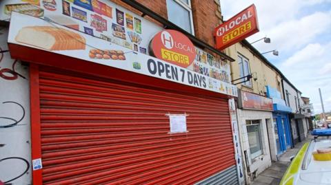 A landscape photo of the outside of H Local Store. The red shutters are pulled down and there is a white paper sign stuck on the front. Above the shutters is a shops frontage with printed items of food and drink that the shop sells