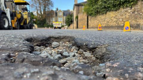 A pothole in the middle of a residential road. Rubble has been exposed due to the surface layer breaking off. The road has industrial machinery parked nearby and is lined with yellow cones.