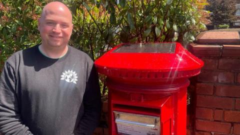 A bald man in a black jumper stands to the left of a red postbox. The postbox had a solar panel on top, a letter slot and a metal pull down drawer. Behind them is a hedge and brick pillar to the right.