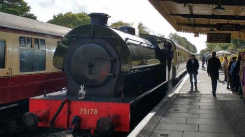 A black steam train waiting at a platform. There are people on the platform to the right of the train. To the left is a train carriage. The sky is cloudy. 