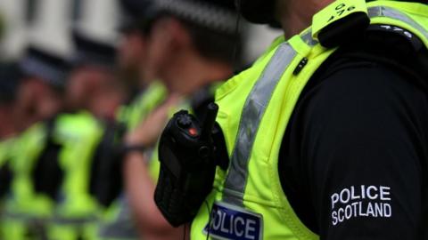 A row of police officers with high vis yellow vests on, and walkie-talkies clipped to their uniform. One of the sleeves has Police Scotland written on it. 
