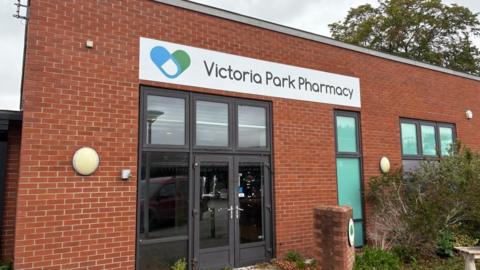 The photo shows a rectangular, one storey red brick building. A large sign above a double glass door reads "Victoria Park Pharmacy". No light comes from inside, with an overcast sky above.