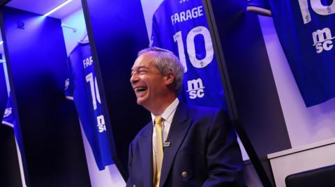 Nigel Farage in a navy suit and a yellow tie sits in Ipswich Town's home dressing room surrounded by personalised "FARAGE 10" Ipswich shirts. He is smiling and laughing as the picture is taken.