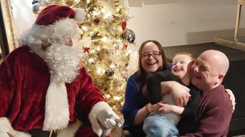 A figure dressed as Father Christmas leans towards Melanie Wallwork and her partner holding their son Jake who is smiling, with the group sat in front of a lit Christmas tree indoors. 